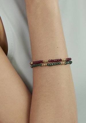 Two beaded bracelets with gold accents, one dark red and one dark green, worn on a slightly bent bare arm against a light background.