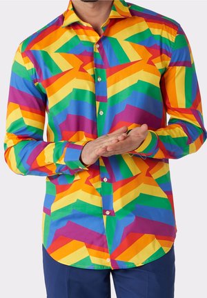 Mann trägt ein Hemd mit einem mehrfarbigen geometrischen Muster in Regenbogenfarben, hält die Hände vor sich verschränkt, vor einem schlichten hellen Hintergrund.