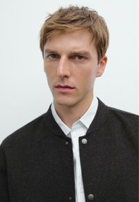 Black bomber jacket with a rounded collar, buttoned front, worn over a white shirt with thin vertical stripes. Neutral background.