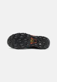 Suola di scarpa da trail running, con battistrada in gomma aggressivo e lugs multidirezionali per una presa migliore, base nera e accenti di branding colorati.