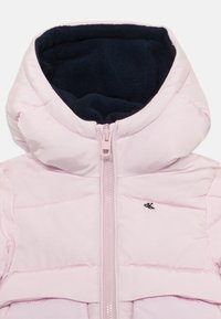 Lys rosa pufferjakke med en marineblå fleece-fôret hette og frontglidelås. Har quiltedetaljer og en liten svart logo på brystet.