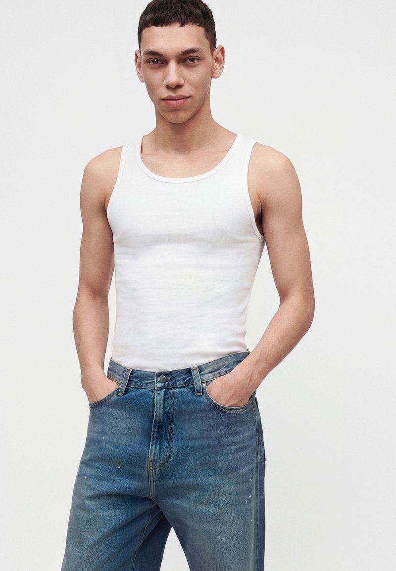 Young man wearing a white tank top and blue jeans with hands in pockets, standing against a plain white background.