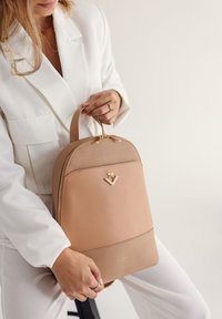 Béžový kožený batoh zaobleného tvaru, se zlatými kovovými prvky a logem na přední straně. Madlo na vrchu s zipy.