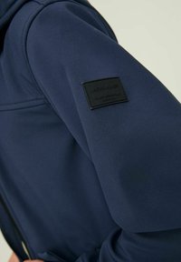 Detalle de chaqueta azul marino con tela suave, capucha ajustable y etiqueta negra con el logo en relieve "JACK & JONES" en la manga.
