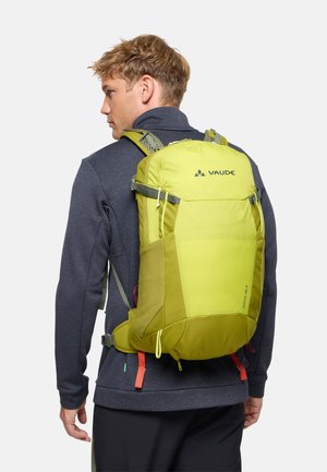 Mann mit dunkler Jacke und hellgrünem Vaude-Rucksack mit mehreren Riemen, von hinten vor einfarbigem Hintergrund gesehen.