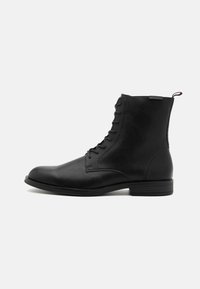 Bota de tobillo de cuero negro con Punta redonda, parte superior texturizada, cordones laterales y una pestaña en el talón. Suela plana con un tread sutil.