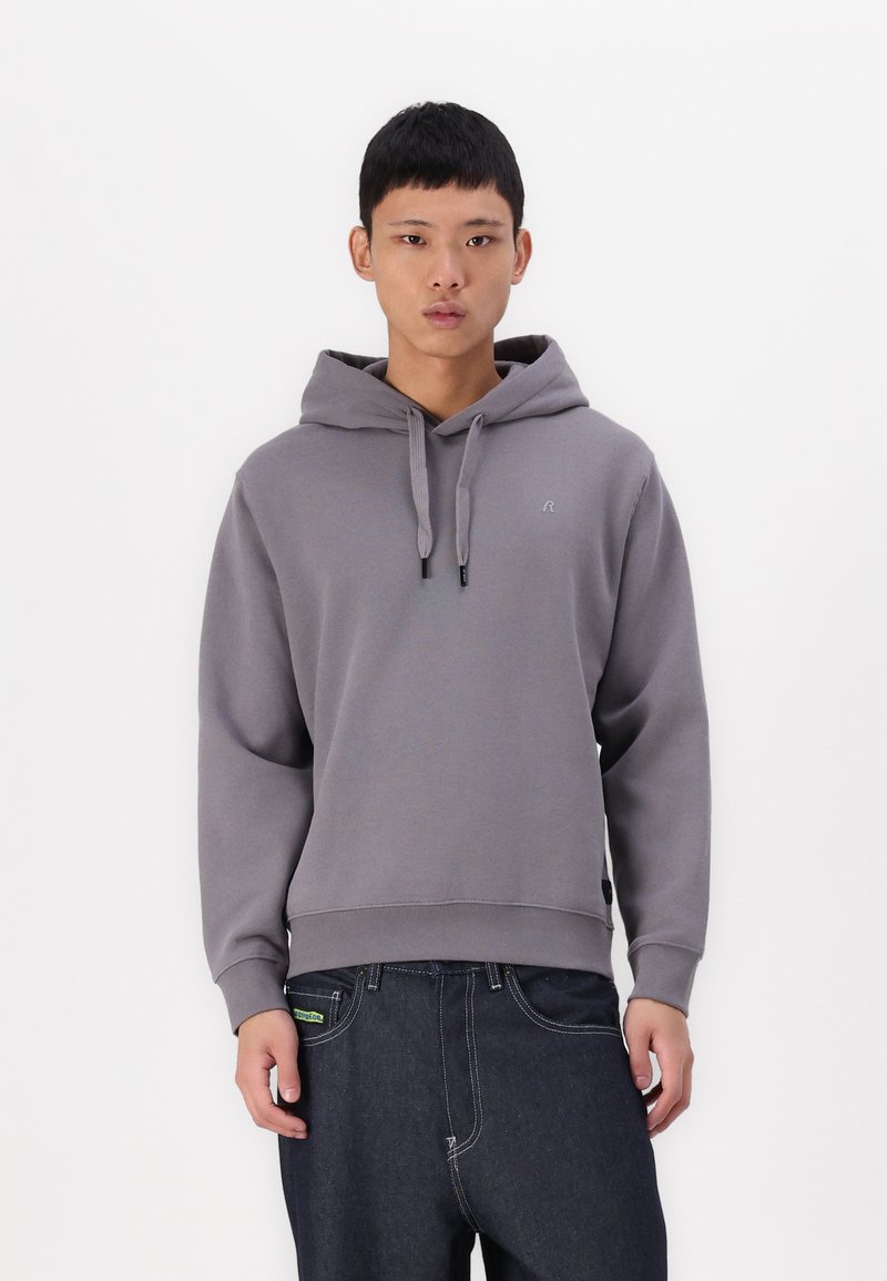 Sudadera gris con capucha y cordones, con puños y dobladillo de canalé. Presenta un pequeño logo en el pecho. Combinada con jeans de mezclilla oscura.
