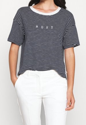 Femme portant un T-shirt rayé bleu marine et blanc avec le texte « ROXY » et un pantalon blanc, mains dans les poches, sur un fond uni.