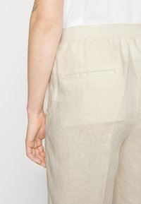 Béžové lněné kalhoty s jednou zadní kapsou, s jemnou strukturou a měkkým, lehkým designem, nošené s bílým topem.