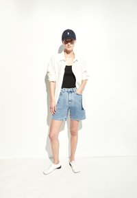 Jeanshorts mit fransigem Saum, ein schwarzes Tanktop, ein weißes Hemd und eine marineblaue Cap, kombiniert mit weißen Schuhen.