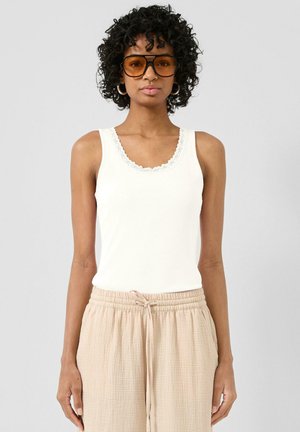 Frau mit lockigem Haar, die große bernsteinfarbene Sonnenbrillen, ein weißes Top mit Spitzenbesatz und beige Hose mit Kordelzug trägt, stehend vor einem einfachen Hintergrund.