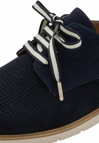 Chaussure en daim bleu marine avec un design perforé, des lacets rayés blancs et bleu marine contrastants, et une semelle en caoutchouc beige. Comprend une étiquette avec un accent métallique.