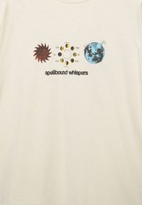 Crème kleurige katoenen t-shirt met een zon, maanfases en een blauwe planeet graphic, met de tekst "betoverde fluisteringen" onder de ontwerpen.