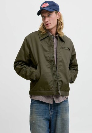 Un jeune homme aux longs cheveux blonds portant une casquette marine, une veste zippée olive, une chemise à carreaux et un jean bleu, se tenant les mains dans les poches.