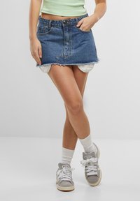 Denim mini skirt with frayed hem, featuring contrast stitching and front pockets. Worn with white ankle socks and grey sneakers.