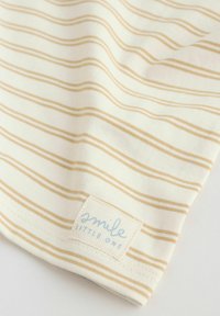 Tissu crème avec de fines rayures horizontales beiges et une petite étiquette cousue portant l'inscription « smile LITTLE ONE » près de l'ourlet.