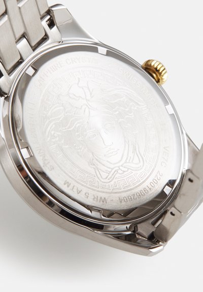 Montre en acier inoxydable argenté avec un dos gravé d'un visage, entouré d'un motif grec. Équipée d'un verre saphir et d'un accent doré.