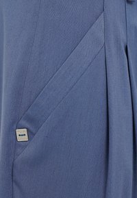 Tessuto blu con una consistenza liscia presenta una piega diagonale e un piccolo cartellino in metallo argentato attaccato all'orlo, aggiungendo un accento sottile.
