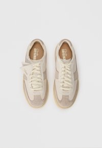 Beige suede sneakers with cream laces, featuring a textured upper and retro design. Soft footbed and rubber sole. Brand logo visible inside.