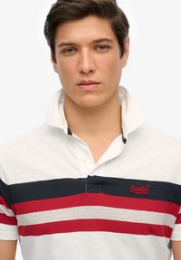 Polo-shirt i hvid med navy og røde vandrette striber. Har en krave, knaplukning og et lille broderet logo på brystet.