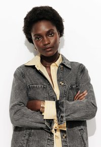 Femme confiante avec des cheveux bouclés naturels, portant des boucles d'oreilles en or, une chemise rayée jaune et une veste en denim grise, les bras croisés.