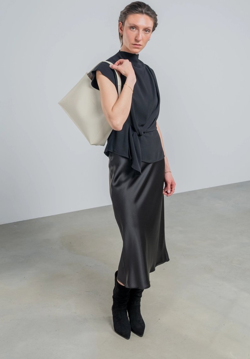 Schwarzes Satin-Kleid kombiniert mit einem figurbetonten schwarzen Oberteil und einer beigen Tragetasche. Das Modell trägt schwarze kniehohe Stiefel. Glatte Texturen und maßgeschneiderte Designs.