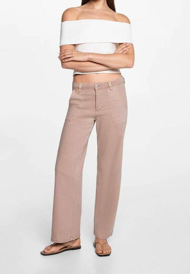 Femme debout, bras croisés, portant un haut blanc épaules dénudées, un pantalon beige taille haute à jambes larges et des sandales plates marron, sur fond blanc.