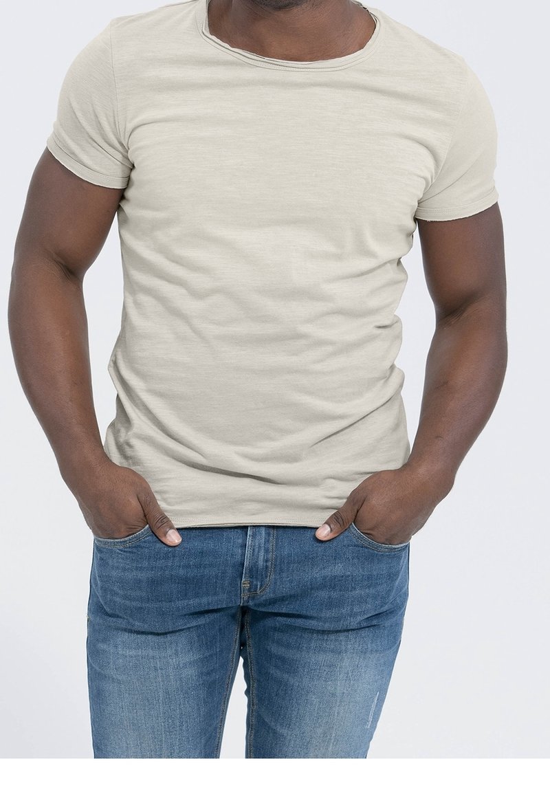 Homme portant un t-shirt uni beige clair à manches courtes et un jean bleu, debout les mains dans les poches devant un fond clair.