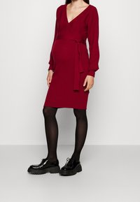 Femme enceinte portant une robe enveloppante rouge à manches longues et longueur genou, des collants noirs et des chaussures à lacets à plateformes noires, se tenant contre un fond uni.