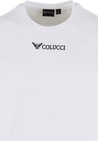 Tricou alb cu guler rotund, având textul „Colucci” negru și un emblema cu aripi imprimată sub linia gâtului, cu eticheta de dimensiune arătând „mare” și fabricat în India.