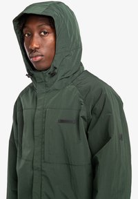 Green hooded jacket with a textured finish, featuring a zippered pocket and branding label on the chest. Adjustable drawstring at the hood.