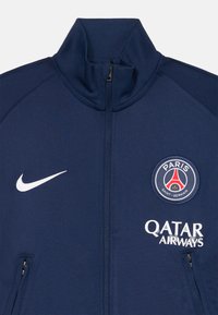 Jūras flotes zipsvārks, izgatavots no gludas auduma, ar augstu apkakli, baltu Nike logo un PSG emblēmu ar sarkaniem, ziliem un zeltītiem akcentiem.