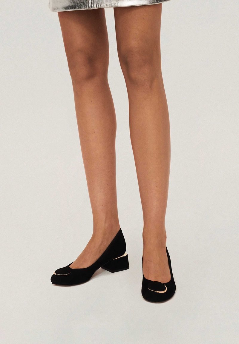 Pumps in pelle scamosciata nera con tacco a blocco, caratterizzati da una punta arrotondata con un accento metallico. Il design include un collo del piede basso per un aspetto elegante.