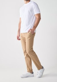 Biała bawełniana koszulka, beżowe chinosy o prostym kroju oraz białe trampki z granatowymi akcentami, stojące na jednolitym tle.