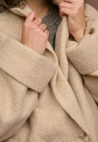 Manteau en mélange de laine beige avec une finition texturée, grands revers et fermeture à un bouton. Des mains saisissent le col, mettant en avant la douceur.