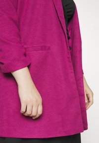 Fuchsia geweven blazer met een enkele knoop, notch-uitsparingen en een zak; gedeeltelijk getoond op de arm van een model. Zwarte top eronder.