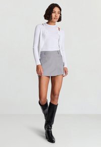 Weißer, gerippter Cardigan mit Knöpfen, kombiniert mit einem grauen, plissierten Minirock und kniehohen schwarzen Lederstiefeln mit runden Verzierungen.