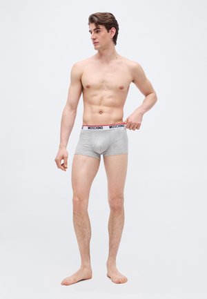 Νέος άνδρας που στέκεται ξυπόλητος φορώντας γκρι εσώρουχα Moschino boxer, κρατώντας τη ζώνη και κοιτάζοντας αριστερά, πάνω σε απλό ανοιχτό φόντο.