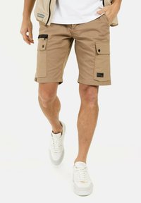 Beige cargoshorts met meerdere zakken en een zwarte ritsdetail, gecombineerd met witte sneakers. De shorts hebben een omgeslagen zoom en een stoffen label.