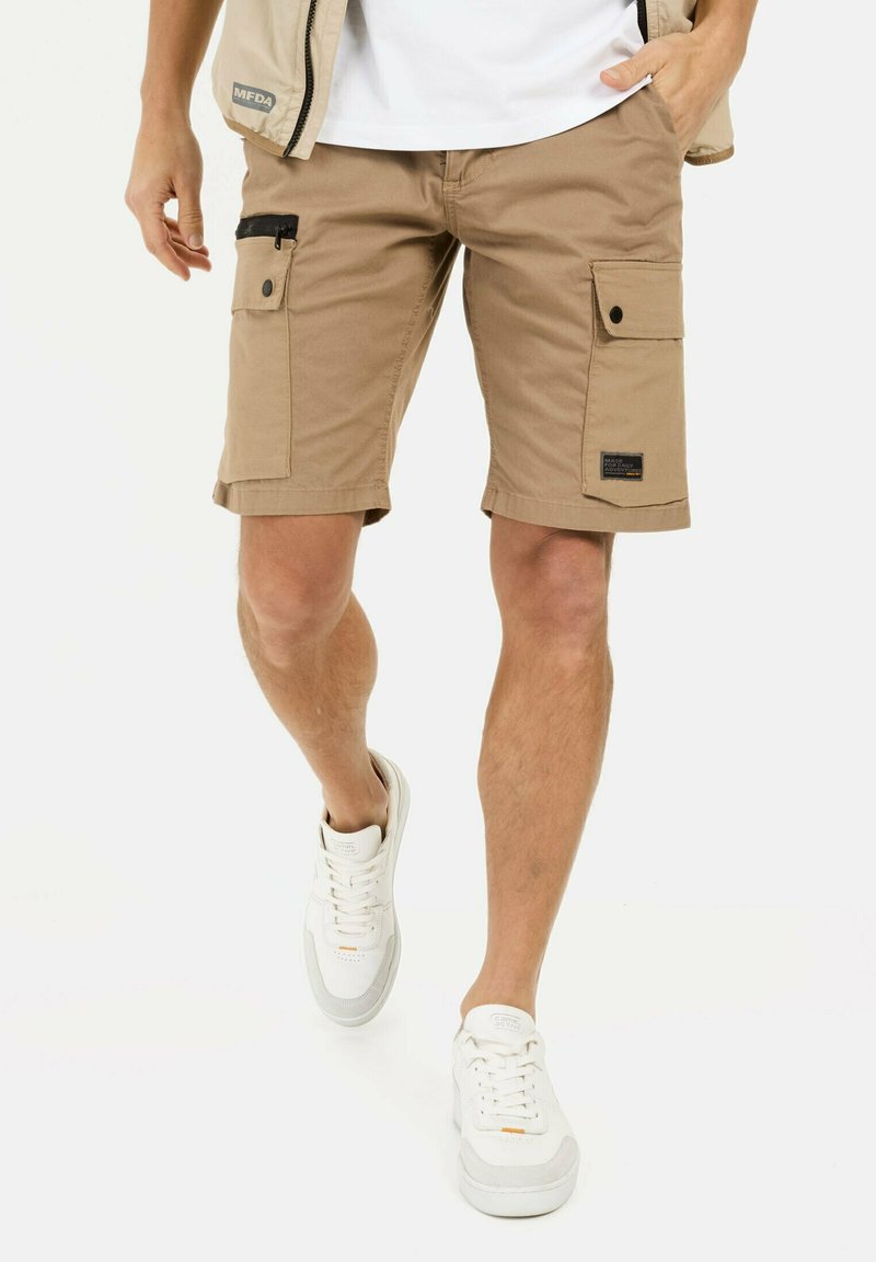 Beige cargoshorts met meerdere zakken en een zwarte ritsdetail, gecombineerd met witte sneakers. De shorts hebben een omgeslagen zoom en een stoffen label.
