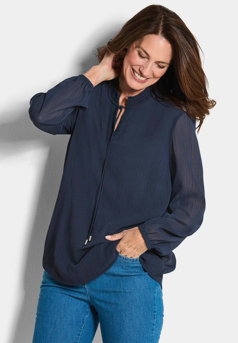Dunkelblaue Bluse mit einem Schlitz-Ausschnitt, langen durchsichtigen Ärmeln und plissierter Textur; kombiniert mit hellblauen Jeans.