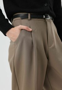 Pantalon beige à coupe ajustée, avec une jambe droite, des poches latérales et une ceinture noire. Le tissu a une texture lisse et structurée.