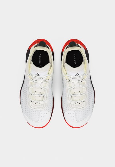 Par belih športnih copatov z luknjicami na straneh, črnimi notranjimi podplati in rdečimi poudarki na podplatu in peti, videnih od zgoraj.