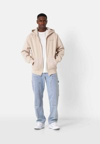 Beige Zip-Hoodie mit Fronttasche, getragen über einem weißen T-Shirt, kombiniert mit hellblauen Cargo-Hosen und weißen Sneakers.