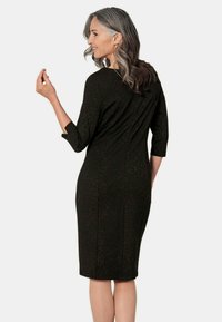 Robe noire à manches trois-quarts, silhouette ajustée, et tissu texturé avec des éclats dorés, se terminant juste en dessous du genou.