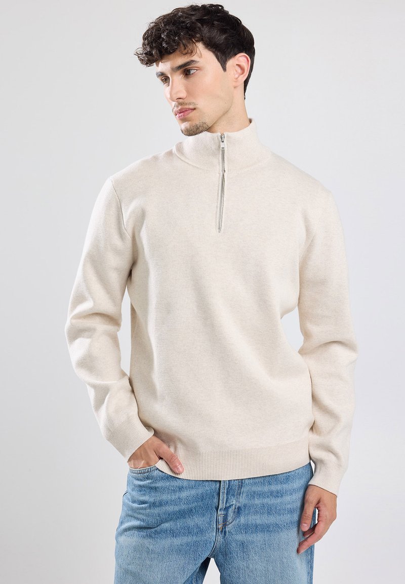 Light beige zip-neck sweater made of soft fabric, featuring long sleeves and a ribbed hem. Worn with blue jeans for a casual look.