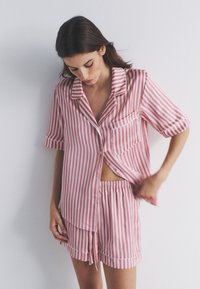 Vrouw die een roze-wit verticaal gestreept kort mouw overhemd met knopen en bijpassende korte broek aanpast tegen een effen muur.