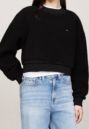 Personne portant un sweat-shirt court noir et duveteux par-dessus une chemise blanche et un jean taille haute bleu clair, main dans la poche.