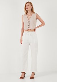 Chaleco de lino beige corto con cinco botones marrones, combinado con pantalones anchos blancos con textura y cordón. Sandalias de tacón marrones completan el look.