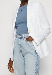Blazer blanco de lino con botones de madera claros, usado sobre una camisa de rayas azul y chaqueta de mezclilla con lavado claro. Jeans rectos con textura azul desvanecida.